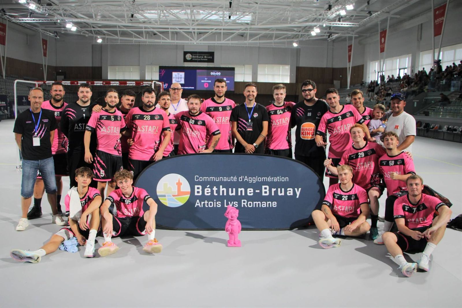 Le groupe seniors en maillot rose et noir avec son staff, dans une grande salle, autour d'un trophée rose