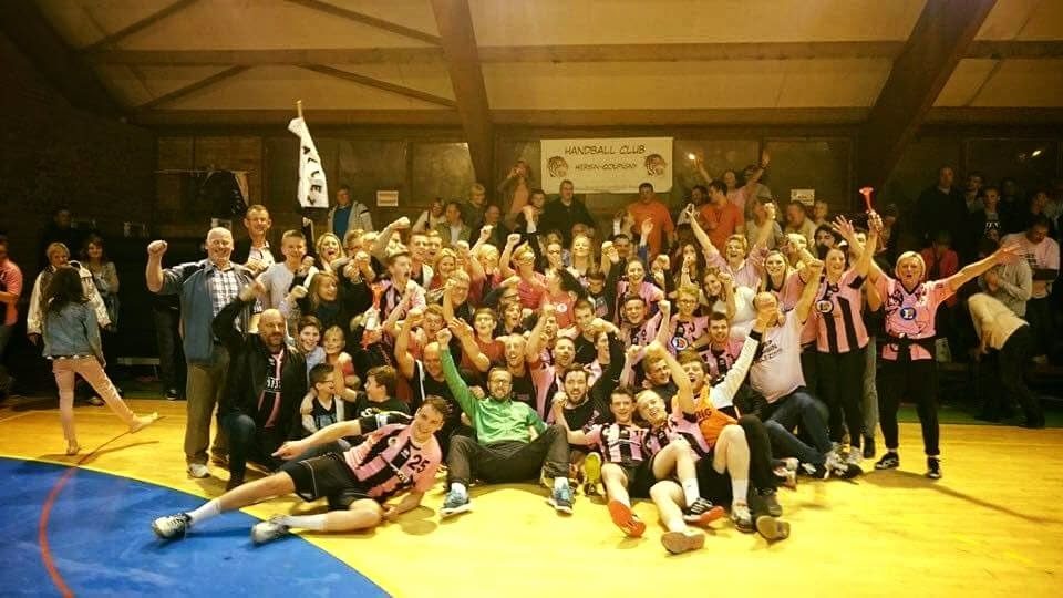 Des dizaines de joueurs en maillot rayé rose et noir, de supporters et de familles, bras levés, fêtent la montée dans la salle de Wahagnies, autour d'un drapeau du club