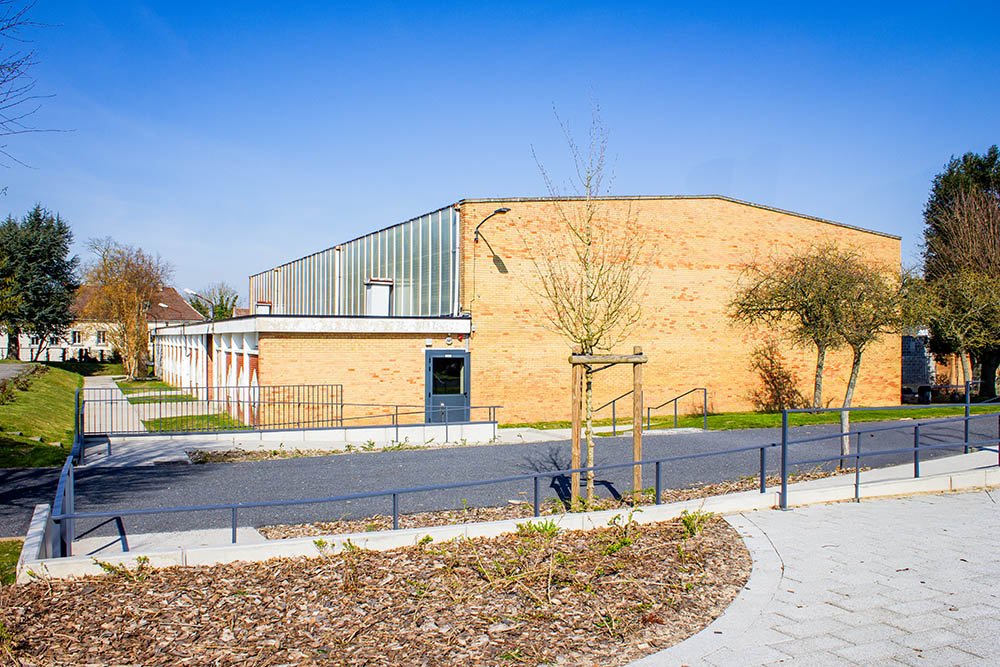 La salle Vivre : un bâtiment en briques claires et bardage translucide, au bout d'une allée